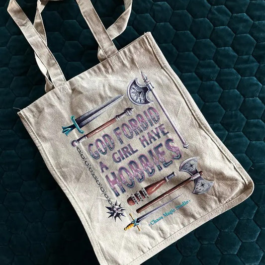 Tote bag with the text "God forbid a girl have hobbies" surrounded by an array of weapons such as a battle axe, mace and dagger