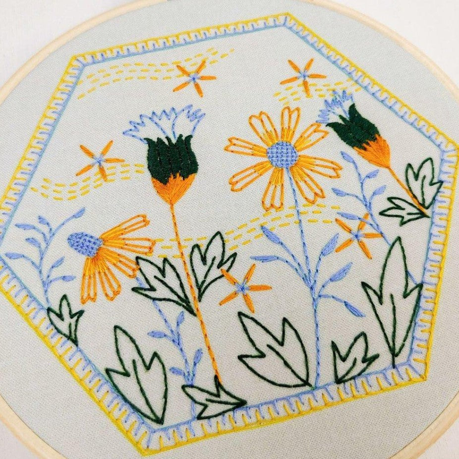 An embroidery kit depicting yellow, blue and black daisies with a hexagonal stitched frame around them.