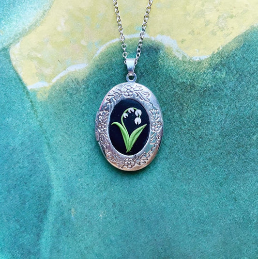 silver locket with flowers etched around the metal edges and a clay sculpted lily of the valley in the center 