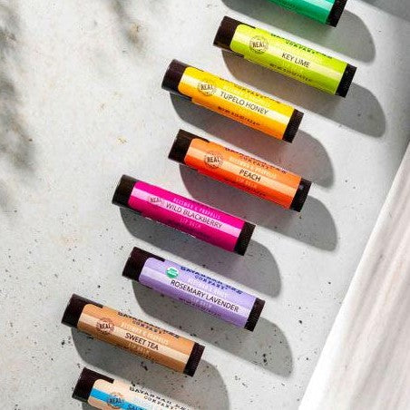 An assortment of Savannah Bee lip balms in the flavors wild blackberry, peach, tupelo honey, key lime, rosemary lavender and sweet tea