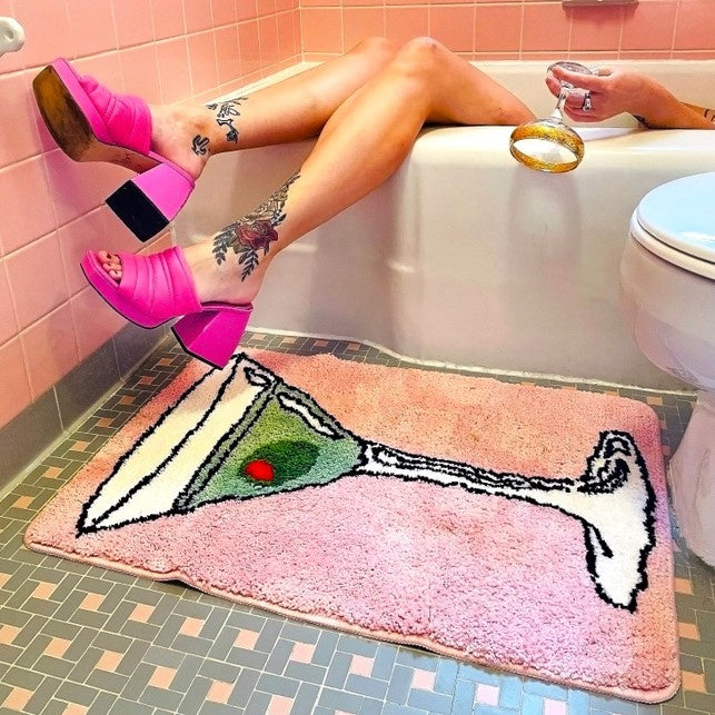 A small plush pink rug being used as a bathmat featuring a martini glass with an olive floating in it.