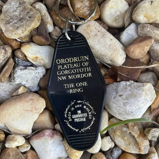 A black plastic keychain with silver sparkly text reading "ORODRUIN PLATEUA OF GORGOTOTH NW MORDOR. THE ONE RING" with a circular sigil logo underneath