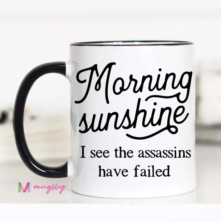 A white mug featuring the words "Morning Sunshine, I see the assassins have failed."