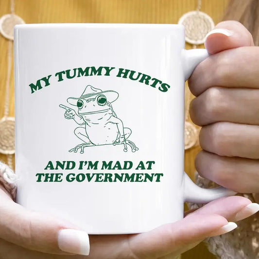 White mug with a green image of a frog in a hat and the words "My tummy hurts and I'm mad at the government"