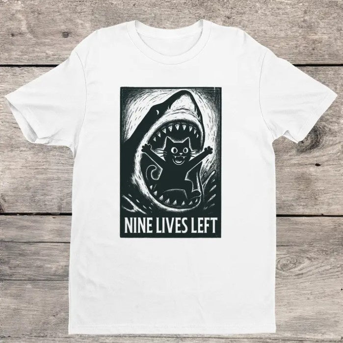 White t-shirt with a black graphic of a smiling cartoon cat jumping out a shark's mouth and text "Nine lives left"