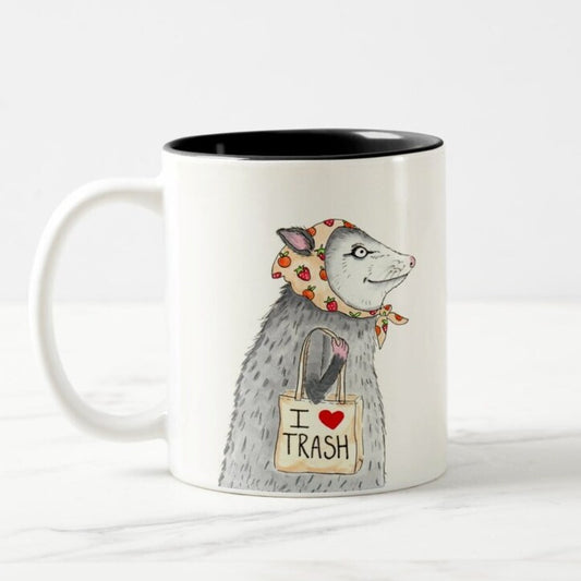 Mug with a black interior and white exterior featuring a design of a possum in a strawberry-printed bandana carrying a tote bag that says "I <3 