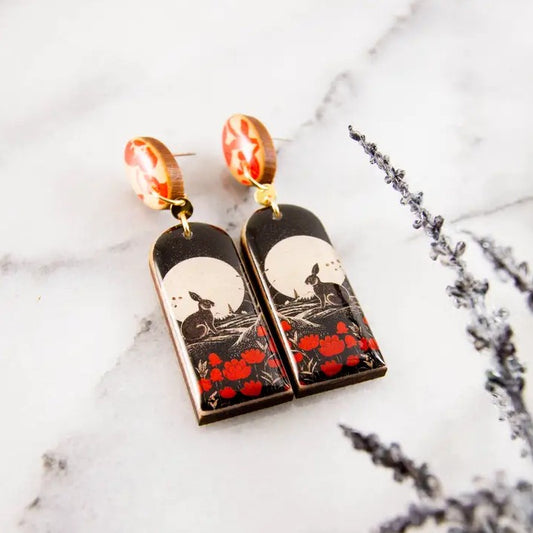 Pair of dangle earrings with a black, white and red image of a rabbit sitting in a poppy field 