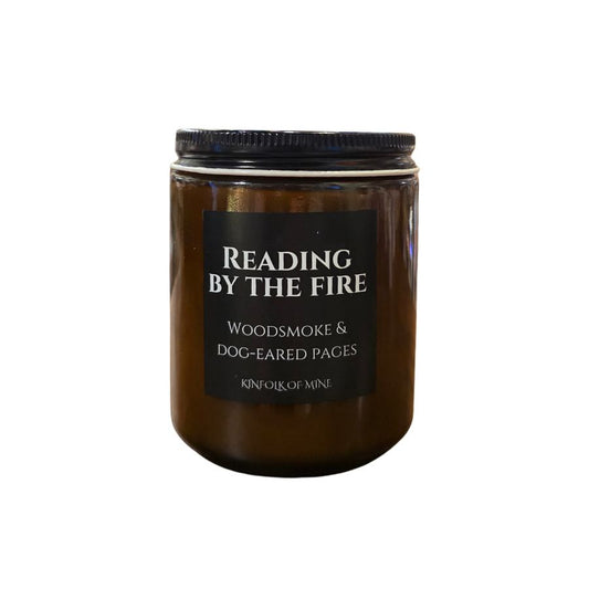 An amber glass candle with a black label that says "Reading by the fire: woodsmoke and dog-eared pages"