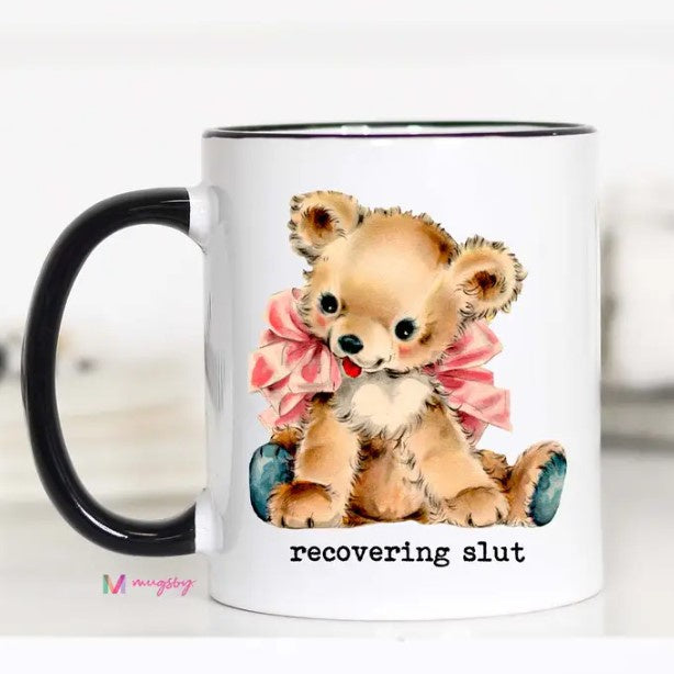 A white mug featuring a retro watercolor illustration of a teddy bear wearing a pink bow and the caption "recovering slut."