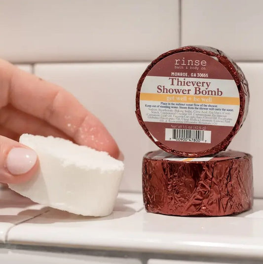 A hand holds a white shower bomb next to two red foil-wrapped Thievery scented shower bombs.
