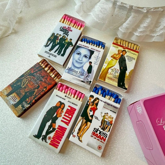 Six novelty matchboxes styled like classic movie matchbooks are arranged on a white textured surface with a lace curtain. The matchboxes feature colorful covers for films including When Harry Met Sally, Pretty Woman, 10 Things I Hate About You, and She's the Man, with various colored match heads visible in the open boxes.