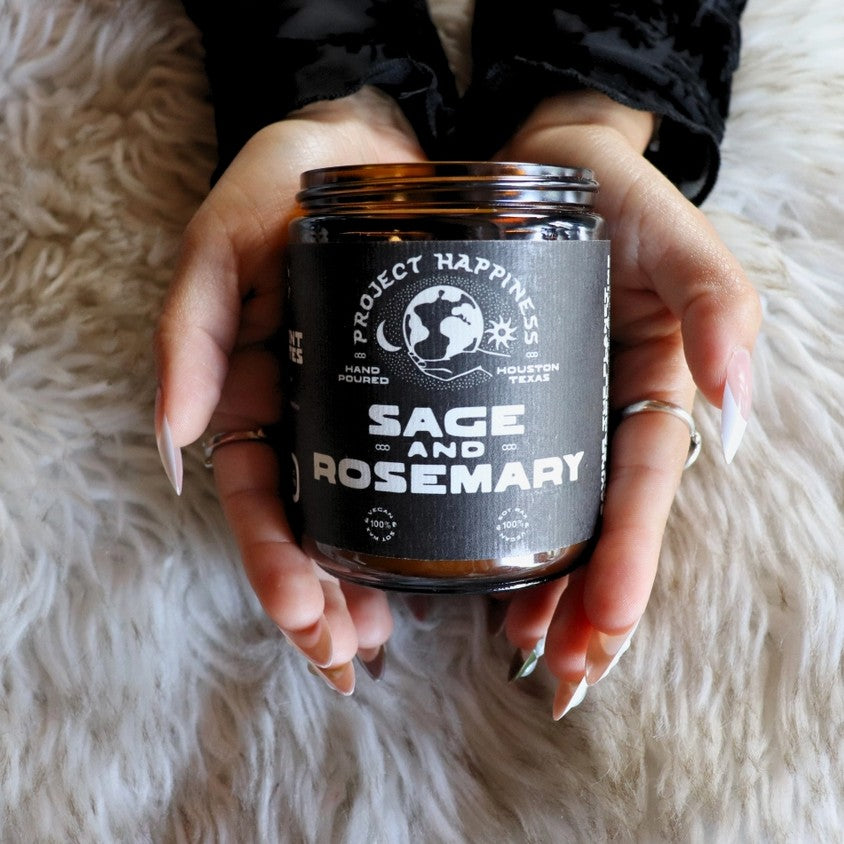 Hands holding a candle in a black jar with the label "Sage and Rosemary"
