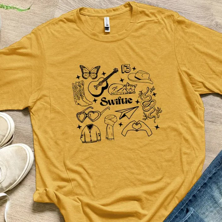 yellow shirt with the word swifties and various symbols relating to the pop artist like the number 13, guitar, and a cat