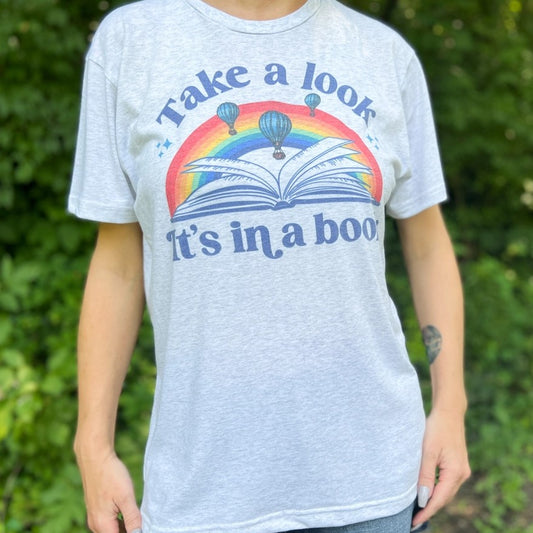 Model Wearing T-shirt that says "Take a look, It's in a book" and in the middle is a rainbow above an open book and air balloons in the sky