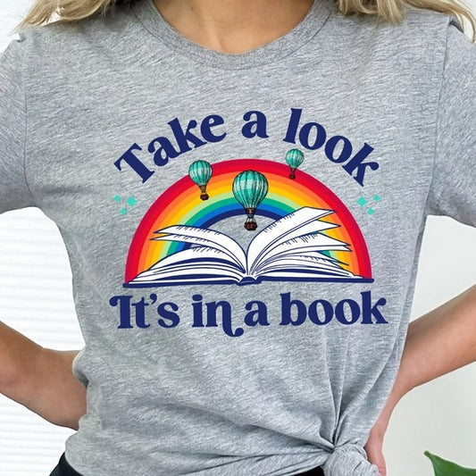 Model Wearing T-shirt that says "Take a look, It's in a book" and in the middle is a rainbow above an open book and air balloons in the sky