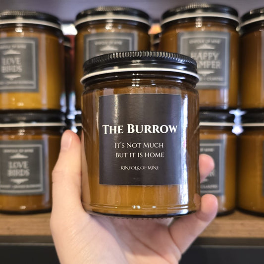 A nine ounce soy wax candle labeled "The Burrow: It's not much but it is home."