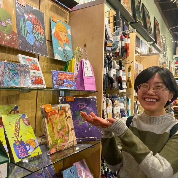 Artist Tiffblot pointing at her art work on a shelf