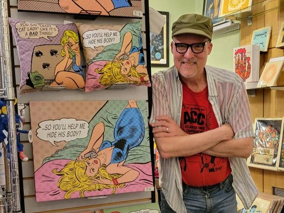 Tom of Bull Moose Publishing standing next to a wall of pillows and prints featuring comedic retro comic book illustrations.