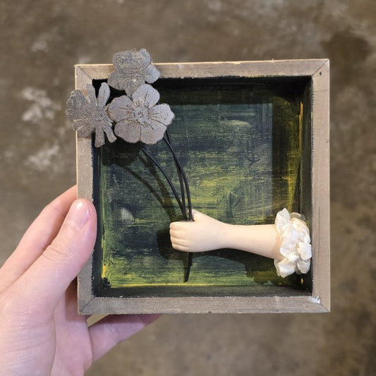 A small shadowbox with a porcelain doll hand holding a bouquet of metal flowers.