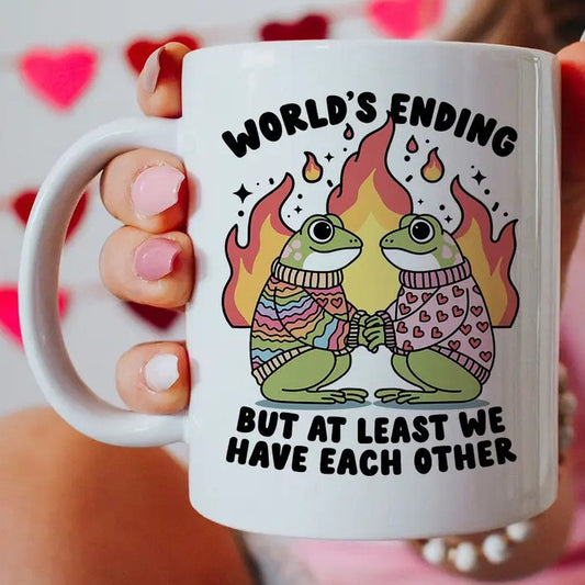 Mug with a graphic of two frogs in sweaters holding hands in front of a fire and text "World's ending but at least we have each other"