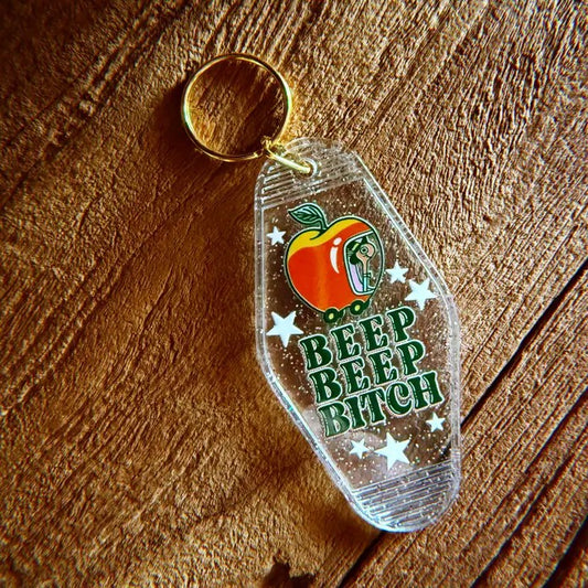 A sparkly translucent plastic keychain with a design of Lowly Worm in an apple car and bold green text "BEEP BEEP BITCH"