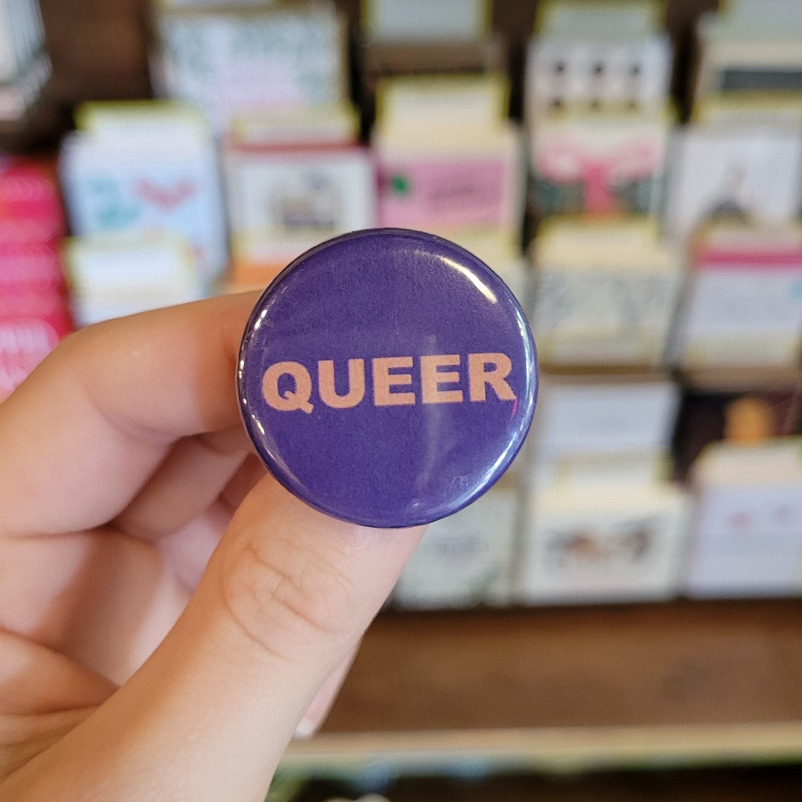 Hand holding pinback button that says Queer